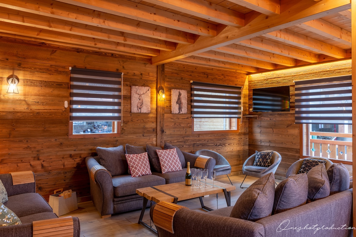 The inviting living area features wooden walls and ceiling beams, with large windows covered by horizontal striped shades. Comfortable seating includes a spacious sofa and armchairs arranged around a wooden coffee table, enhancing the cozy ambiance. A television is mounted on the wall, completing the setting.