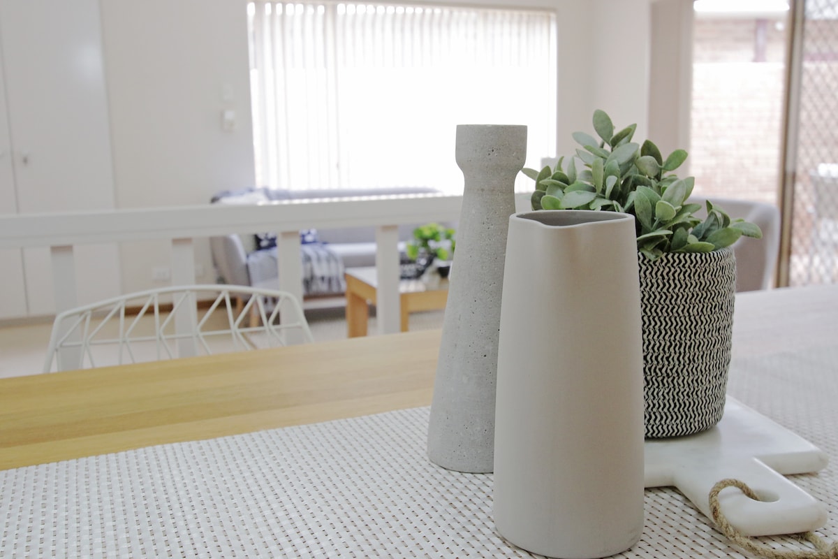 A dining table is adorned with modern ceramic vases and a potted plant, adding a touch of greenery. In the background, the living area is visible through large sliding doors, featuring a comfortable sofa and natural light from the windows.
