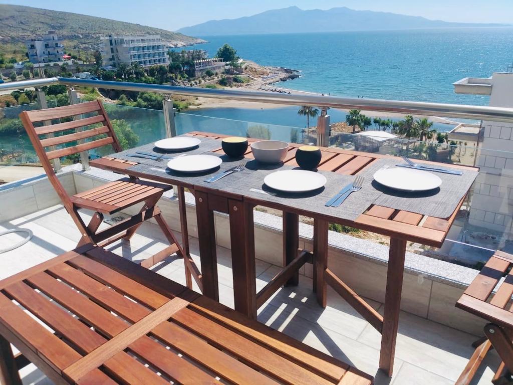 A furnished balcony is presented with a wooden dining table set for four, featuring plates and bowls. The view beyond encompasses the Ionian Sea and its distant mountains, enhancing the tranquil environment.