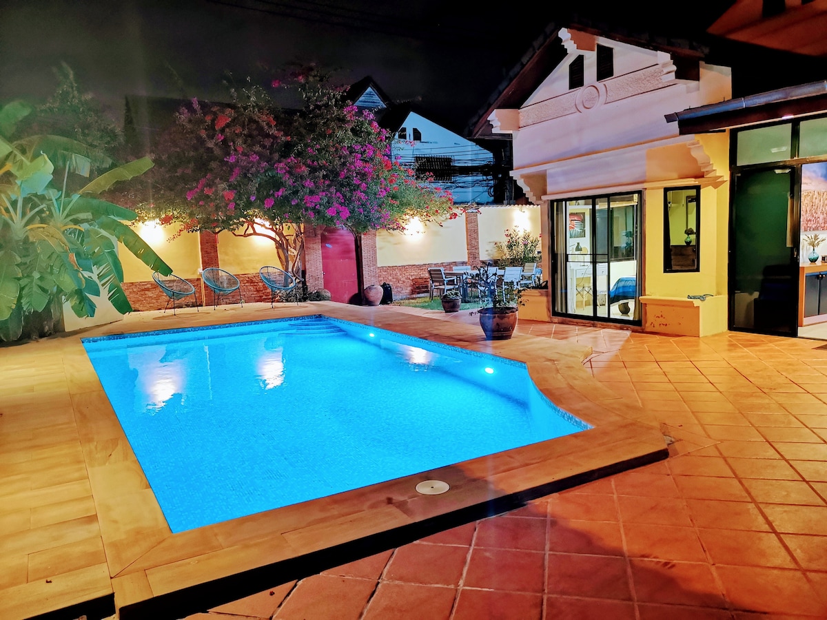 A private salt-water pool glows in soft blue light, surrounded by a tiled patio. Lush greenery and flowering plants frame the area, while a cozy seating arrangement is visible nearby. Floor-to-ceiling glass doors lead into the villa, enhancing the indoor-outdoor flow.
