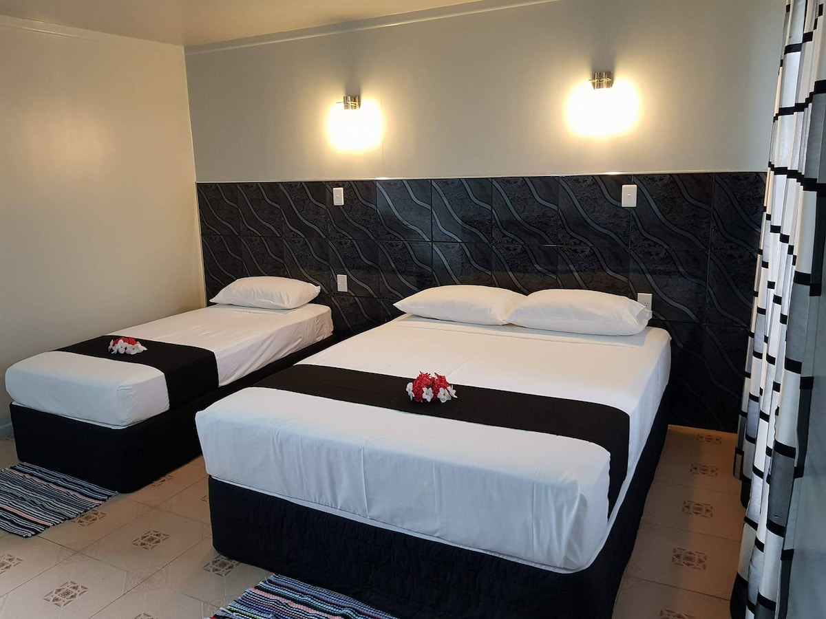 A sleeping area features two spacious beds with crisp white linens and decorative black throws. Soft floral accents are placed on the beds. The walls are adorned with a textured design, complemented by two wall-mounted lights providing gentle illumination.