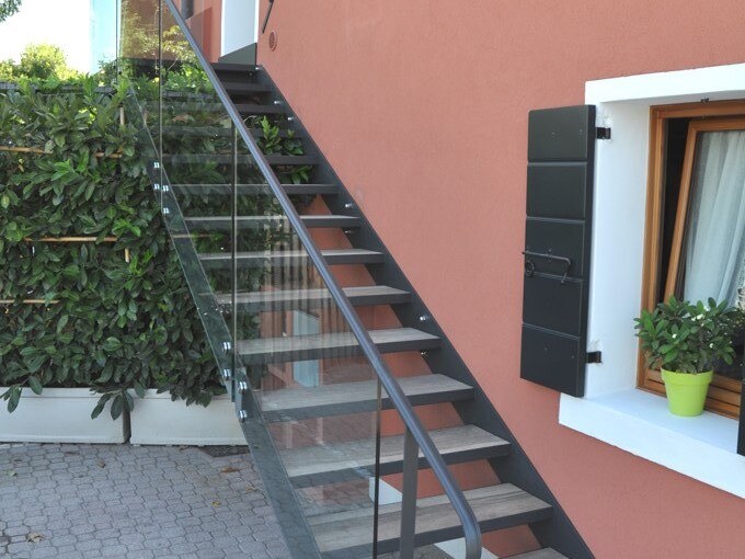 An exterior staircase is seen leading to the upper levels of the building. It features a sleek metal railing and glass panels, with wooden steps offering a contemporary look. Lush green hedges border the area, alongside a window framed in white with a potted plant.