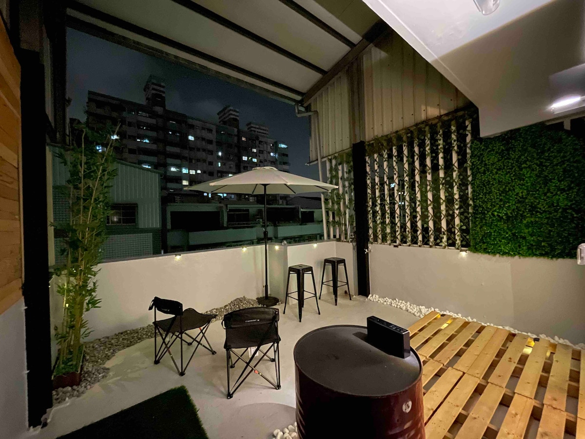 An outdoor terrace features a metal table with an umbrella, surrounded by two folding chairs and two high stools. String lights illuminate the space, and a wooden deck is laid out on one side. A green wall adds a touch of nature to the area.