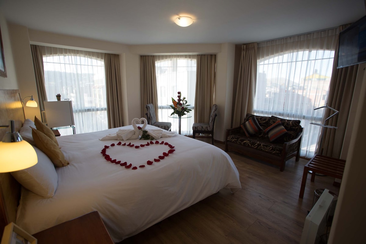 A spacious bedroom features a large bed adorned with heart-shaped arrangement of flower petals. Natural light filters through large, soundproof windows, illuminating the room. A sitting area with comfortable chairs and a decorative sofa complements the modern decor, creating a welcoming atmosphere.