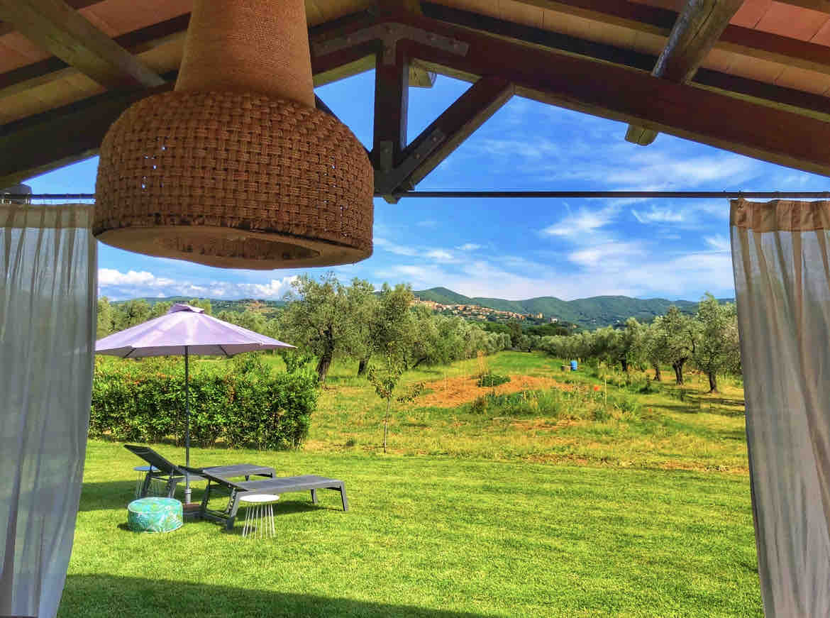 A spacious outdoor area is shown, framed by a wooden roof. Two loungers are positioned on lush green grass, under a shaded umbrella. A decorative woven light fixture hangs above. Rolling hills and olive trees create a serene backdrop against a clear blue sky.