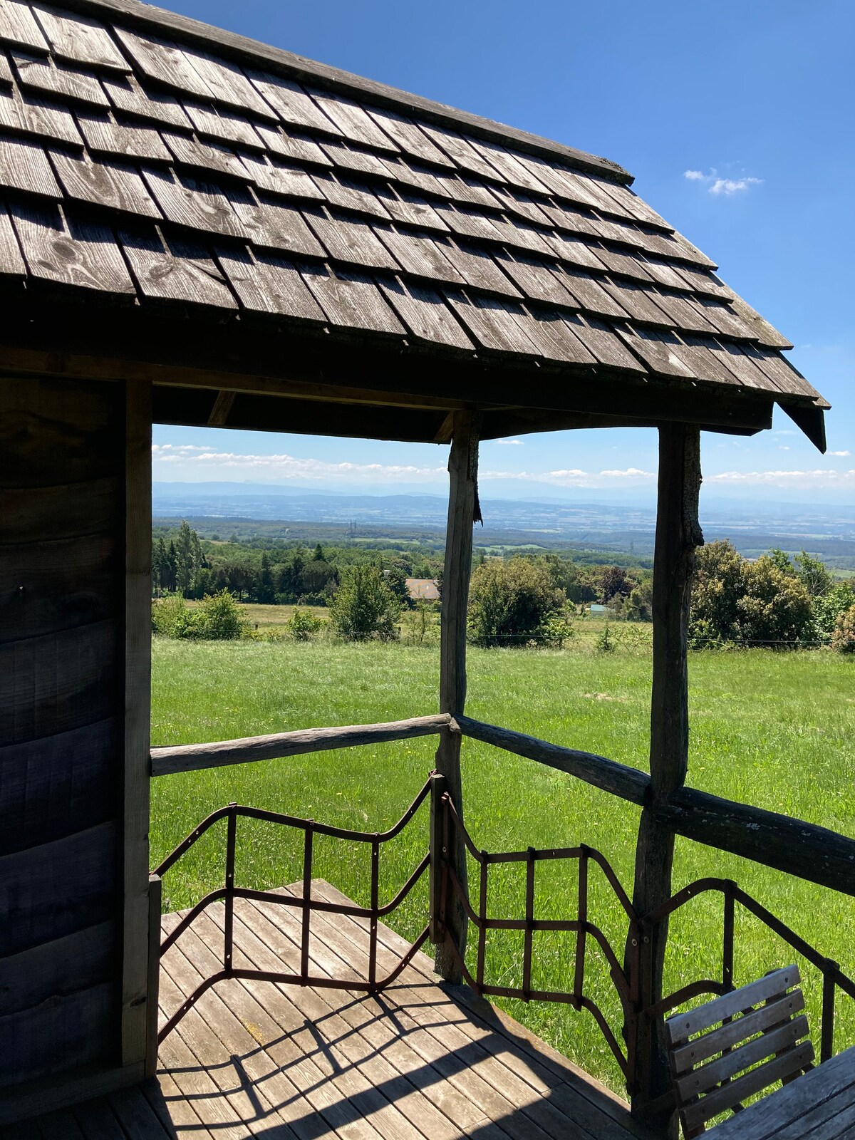 A wooden balcony leads out from the cabin, framed by rustic beams. The expansive view reveals rolling green hills and a distant horizon under a clear blue sky, providing a serene backdrop to the tranquil surroundings.