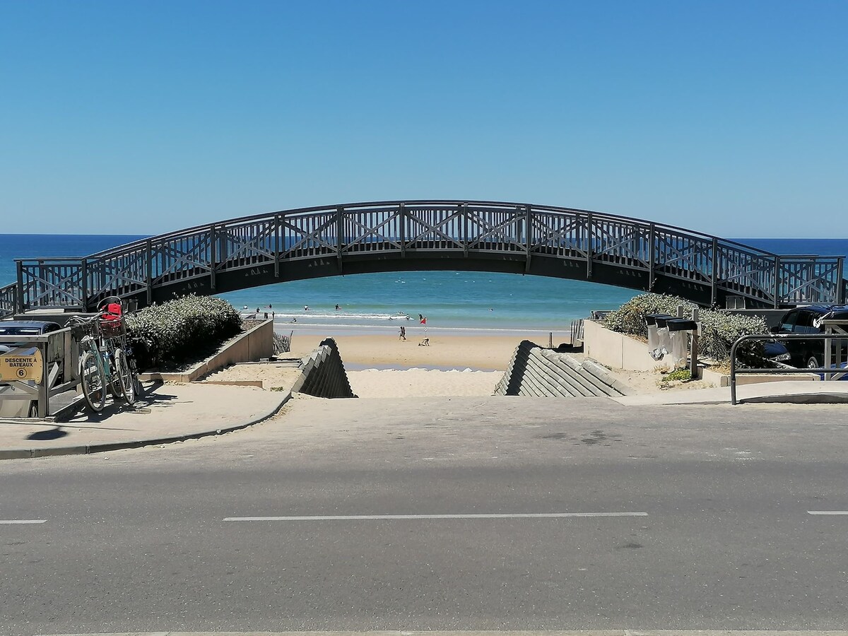 A wooden pedestrian bridge is prominently featured, leading over a sandy path to a beach with visible ocean waves and people enjoying the water. Bicycles are parked nearby, with a clear blue sky overhead enhancing the coastal scene.