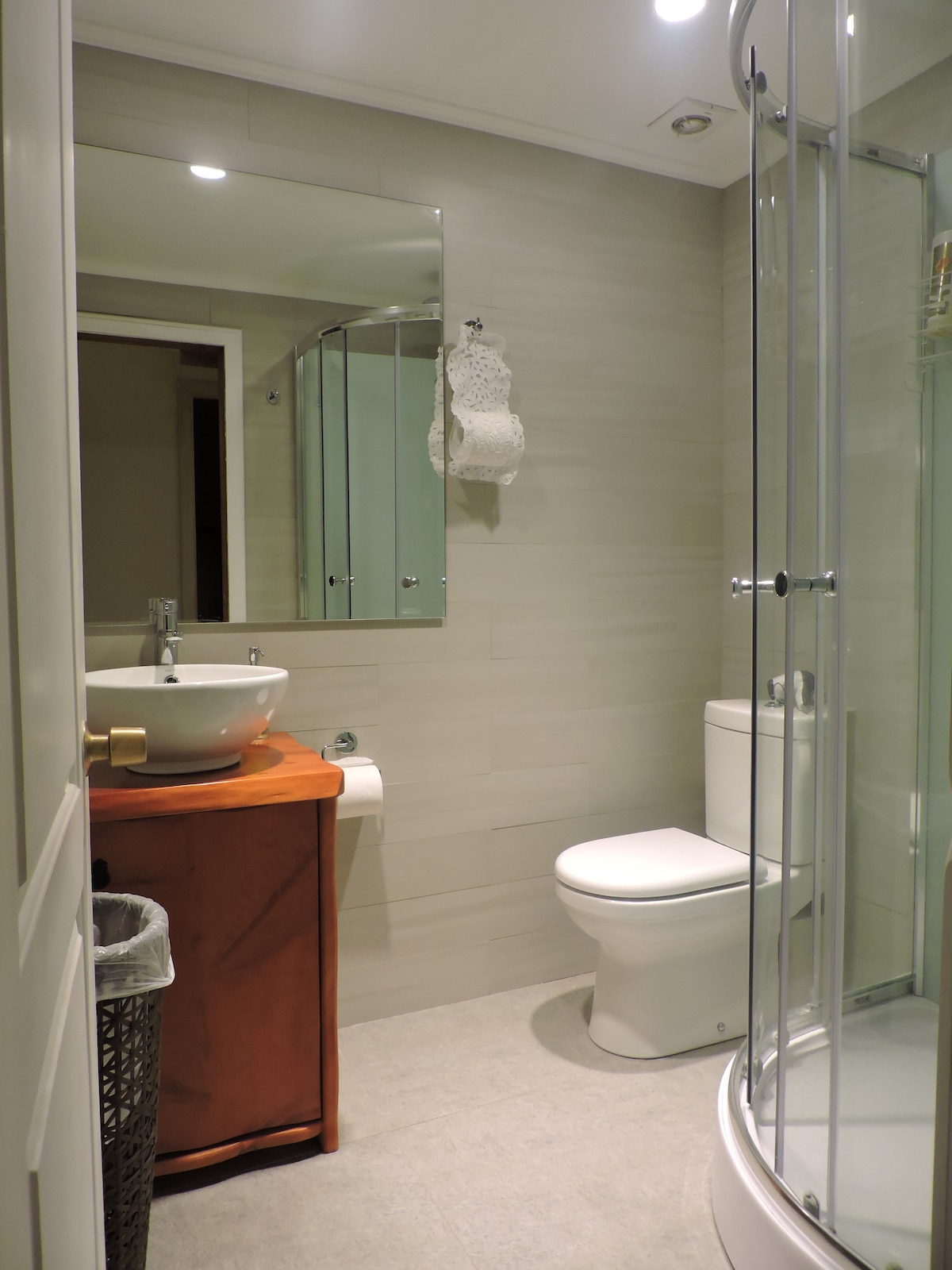 A modern bathroom is presented with neutral-toned walls and sleek fixtures. A round sink sits atop a wooden surface, with a large mirror above. A glass shower enclosure is visible to one side, and a white toilet is positioned next to it. Floor tiles are light in color.