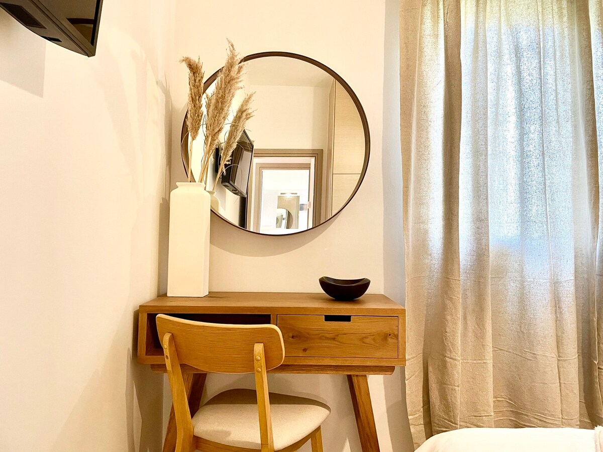 A minimalist workspace is featured, with a wooden desk and a light-colored chair. An oval mirror hangs above the desk, reflecting natural light from a nearby window dressed in soft, sheer curtains. A vase with decorative grasses adds a touch of elegance to the arrangement.