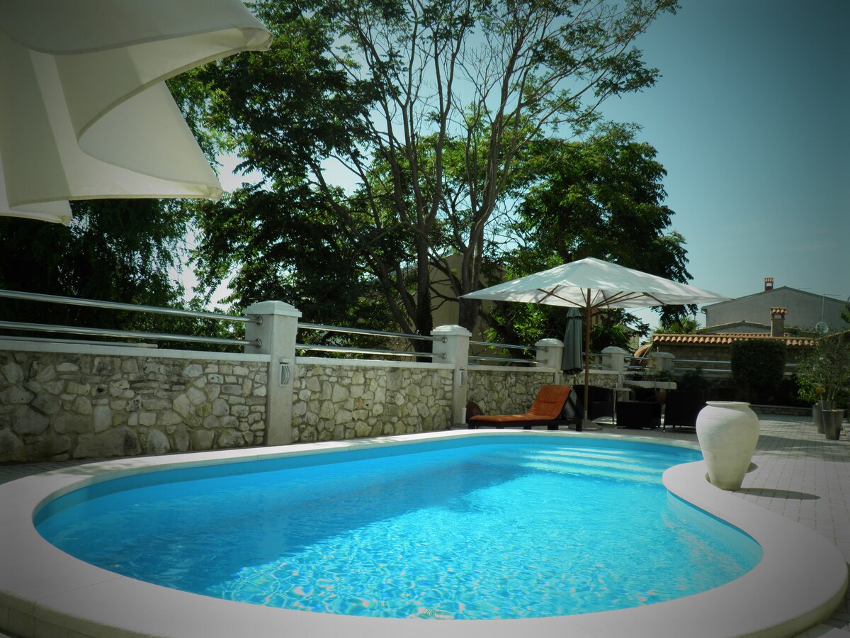 A tranquil swimming pool is surrounded by a paved area, featuring sun loungers and umbrellas for shade. Lush trees and greenery create a serene backdrop, while a stone wall adds a touch of elegance to the outdoor space.
