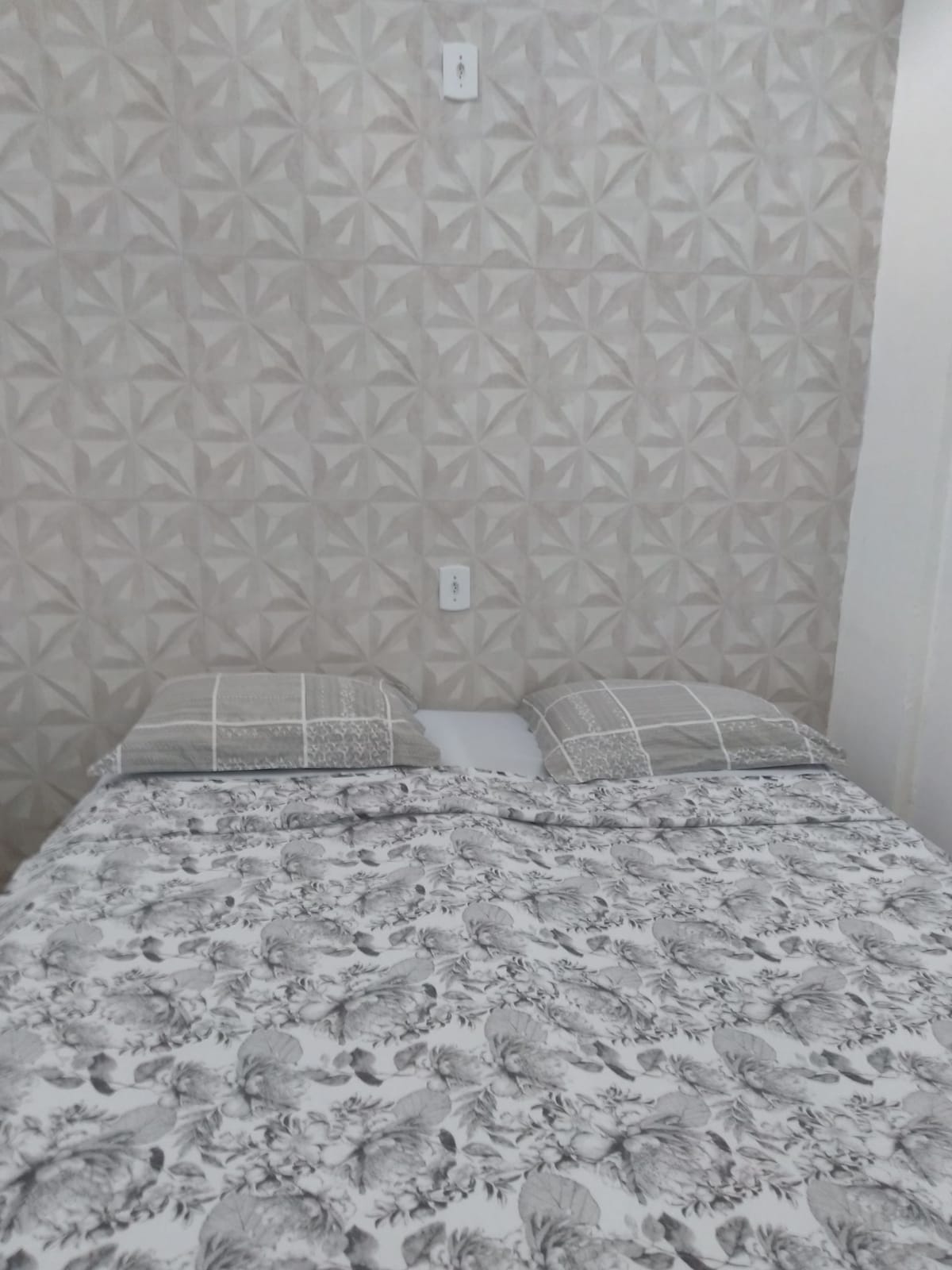 A simple sleeping area features a spacious bed with two pillows placed atop a patterned quilt. The wall behind is decorated with a subtle geometric design in soft tones, creating a calm ambiance.