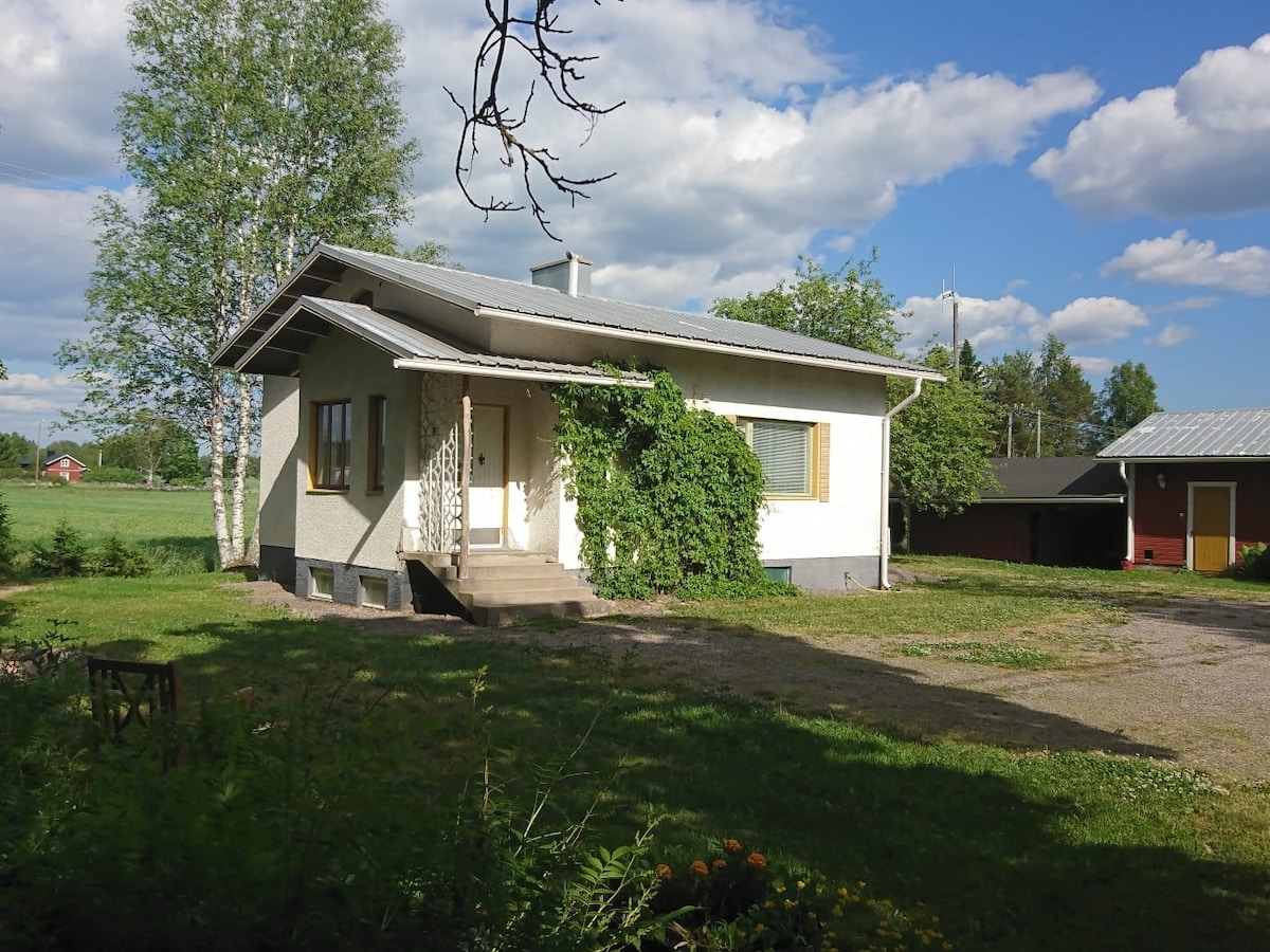 The exterior of the house is shown, featuring a simple design with light-colored walls and a sloping roof. Surrounding greenery and trees provide a natural backdrop. A gravel pathway leads to the entrance, which is framed by a vine-covered trellis. The setting is peaceful and open.