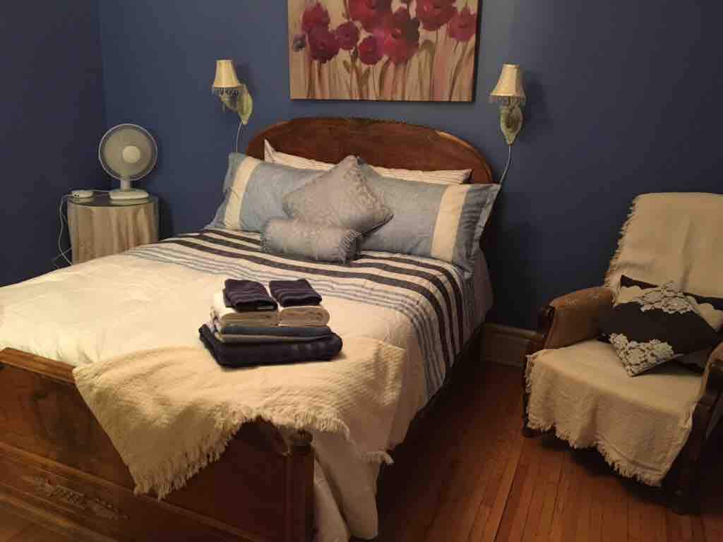 A cozy bedroom is presented with a double bed dressed in soft linens and decorative pillows. A vintage armchair sits adjacent, with a light throw upon it. A floral artwork adorns the wall, and a small fan is placed on a bedside table.