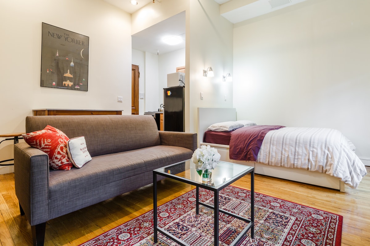 A spacious private studio is shown, featuring a full-size bed and a comfortable sofa arranged around a glass coffee table. A small kitchenette is visible, and warm wooden floors contribute to the overall inviting feel of the space.