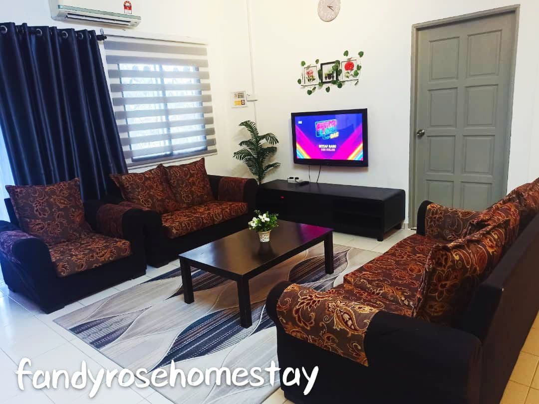 A spacious living area is furnished with a dark-colored sofa set adorned with patterned cushions, complemented by a coffee table at the center. A wall-mounted television displays colorful content, while natural light filters through partially closed window shades. A small plant adds a touch of greenery.