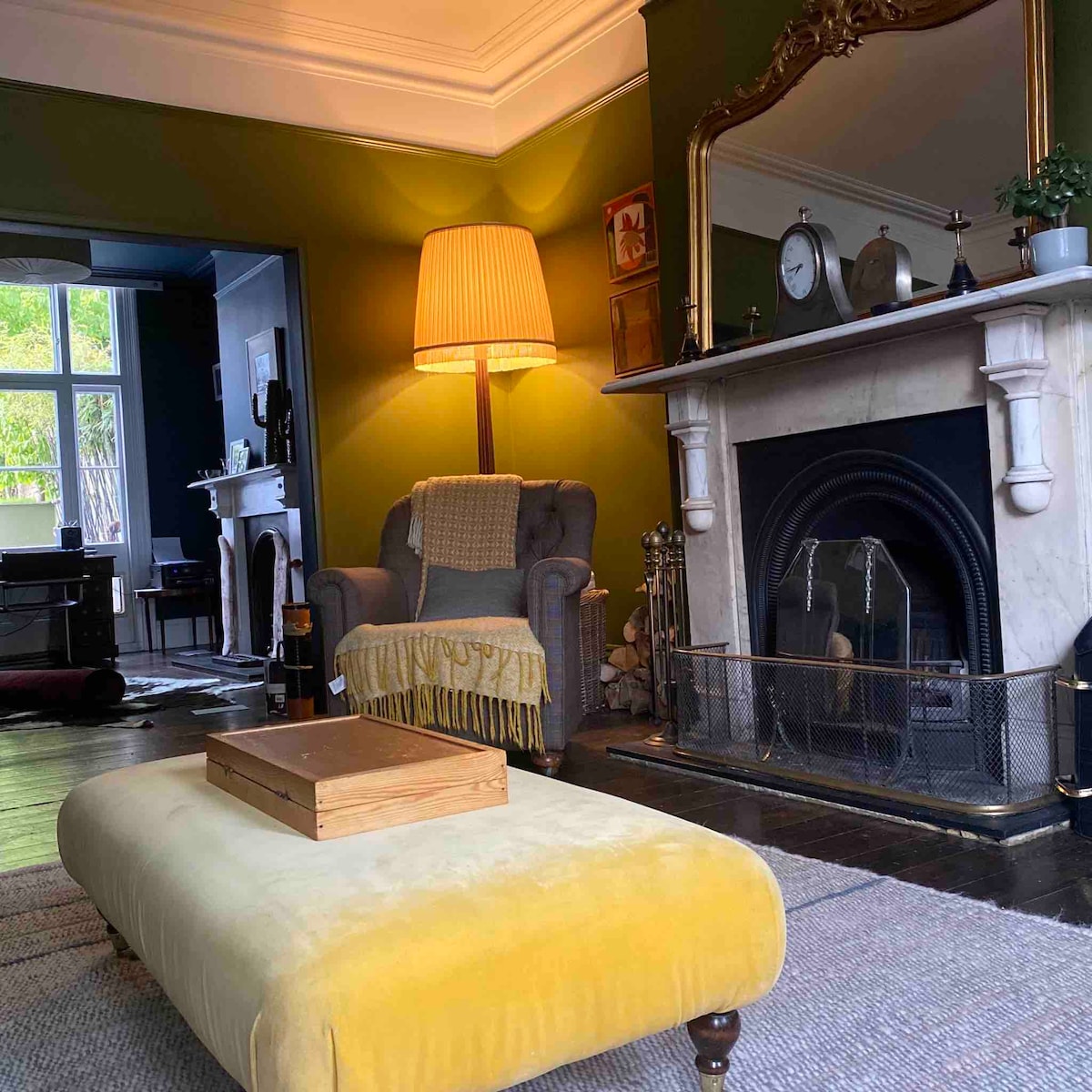 A welcoming sitting room features a vibrant yellow ottoman at its center, flanked by a plush armchair and a large fireplace. Soft light emanates from an elegant lamp, while a decorative mirror and subtle green walls enhance the room's inviting atmosphere.