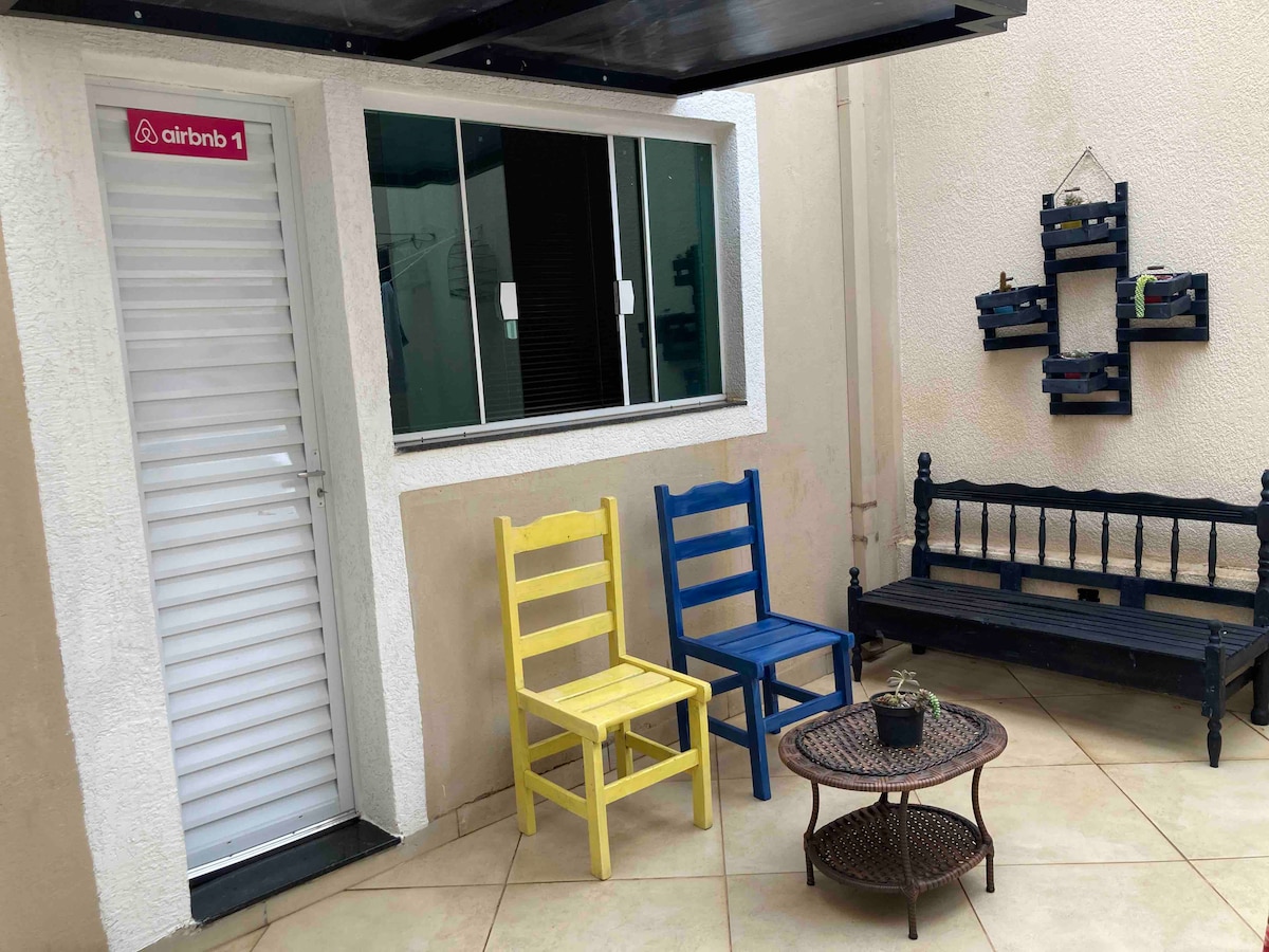 An outdoor patio area is showcased, featuring a mix of colorful chairs in yellow and blue, alongside a black bench. A small round table rests in the center, with a potted plant on top. Natural light illuminates the space, enhancing the inviting atmosphere.