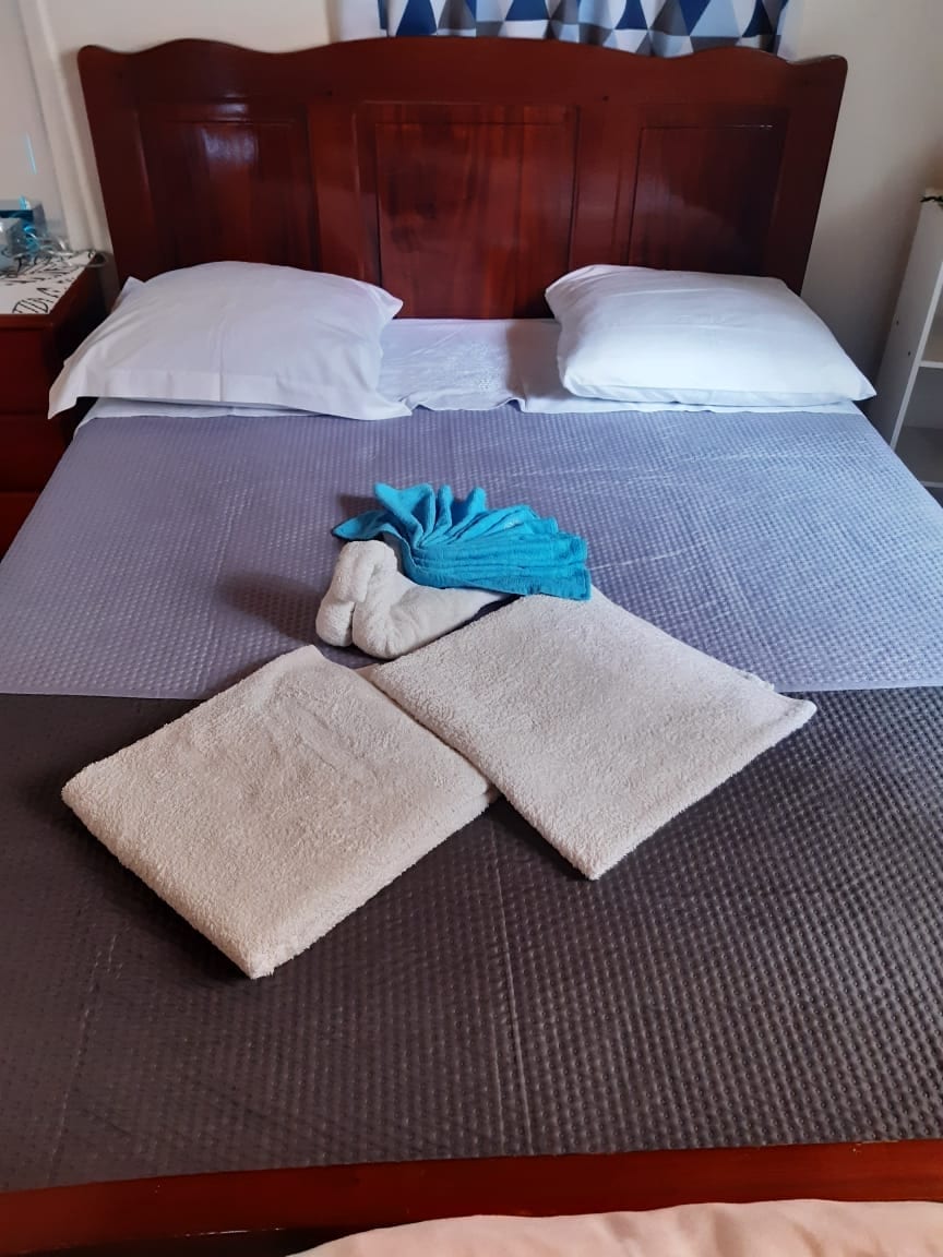 A spacious bed is featured with white pillows and a gray quilted cover. Neatly arranged on the bed are two rolled towels and two larger bath towels in soft, neutral colors. A decorative blue towel adds a touch of color.