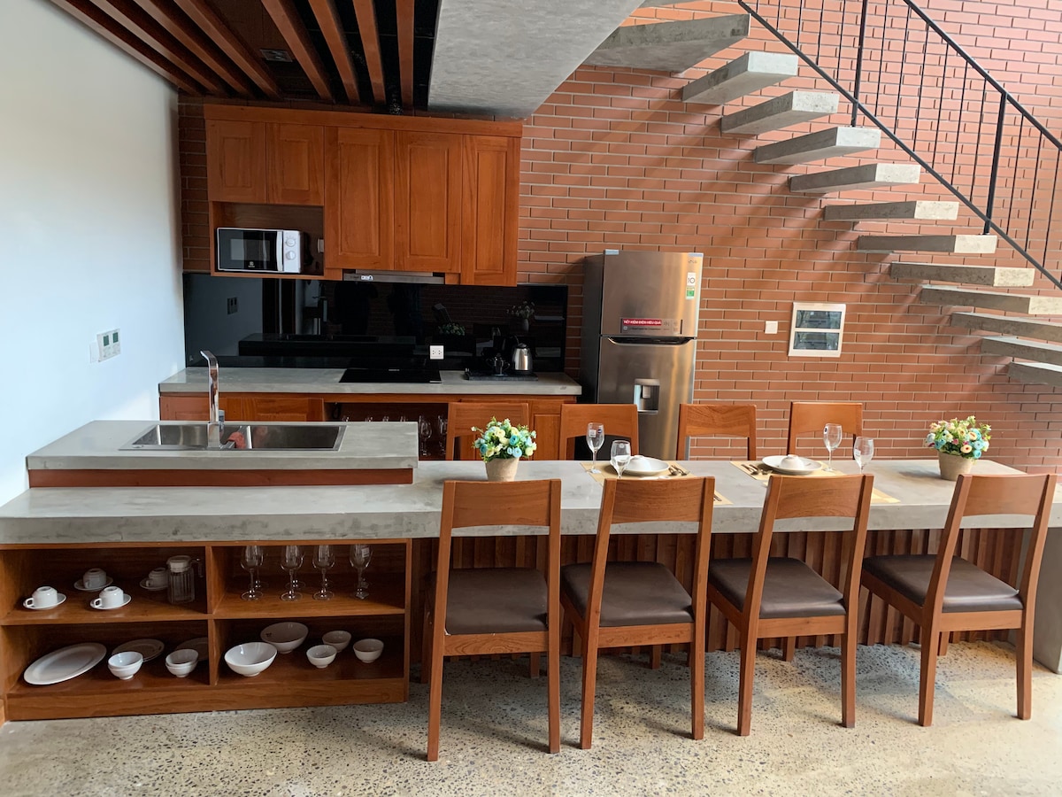 The kitchen area features rich wooden cabinetry and modern appliances, including a refrigerator and microwave. A large island with a sink is complemented by a long table with high-backed chairs, ready for dining. Natural light fills the space, highlighting the warm tones of the materials.