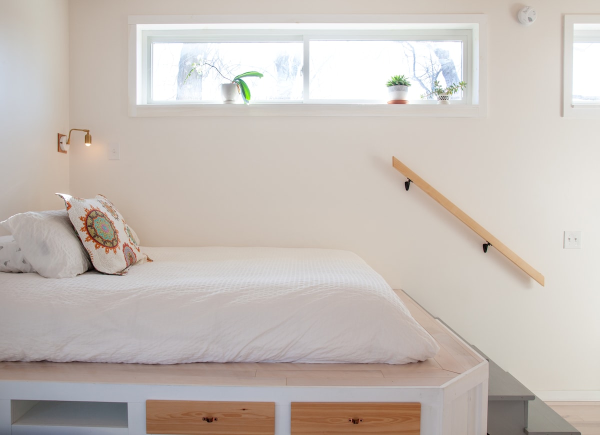 An elevated queen bed is showcased with a white comforter and a decorative pillow. Natural light filters through a large window above, accompanied by small potted plants. A wooden staircase leads to a different area, enhancing the studio's overall spacious feel.