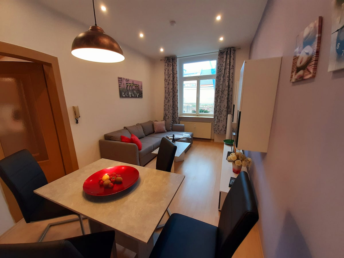 A living area with a gray sofa, decorated with colorful cushions, is accompanied by a dining table set for four. The space is illuminated by natural light streaming through large windows, and floral accents are placed on the table, enhancing the room's inviting atmosphere.