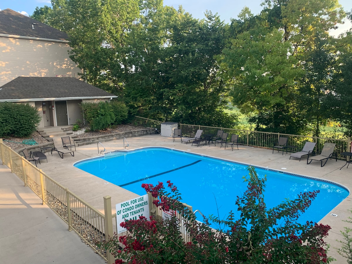 An outdoor pool area is surrounded by trees, featuring a bright blue pool with several lounge chairs arranged around it. A sign indicates the pool is for guest use, and nearby is a well-maintained pathway leading to the entrance of the condo.