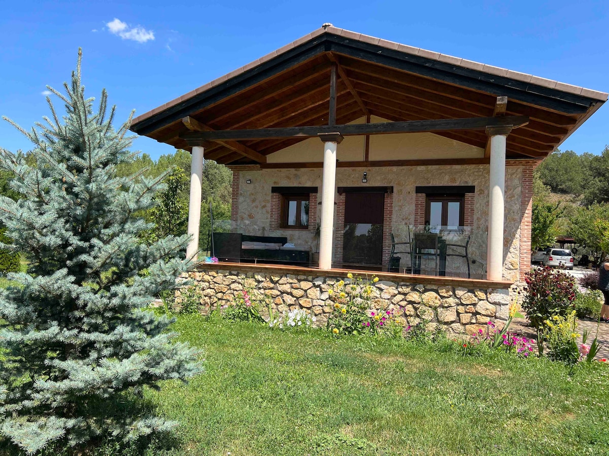 A rustic house features a covered porch supported by white columns, overlooking a well-maintained garden. Vibrant flowers and greenery surround the stone base, while a blue sky with scattered clouds sets a serene backdrop.