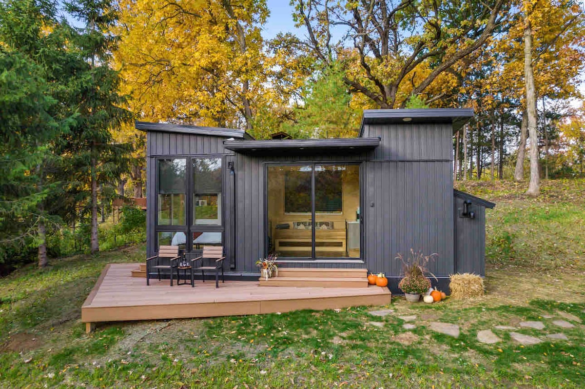 A modern tiny house exterior showcases a sleek black facade with large glass doors, inviting in natural light. A cozy deck features two chairs and seasonal decor, including pumpkins and hay bales. The surrounding landscape includes vibrant autumn foliage and lush greenery.