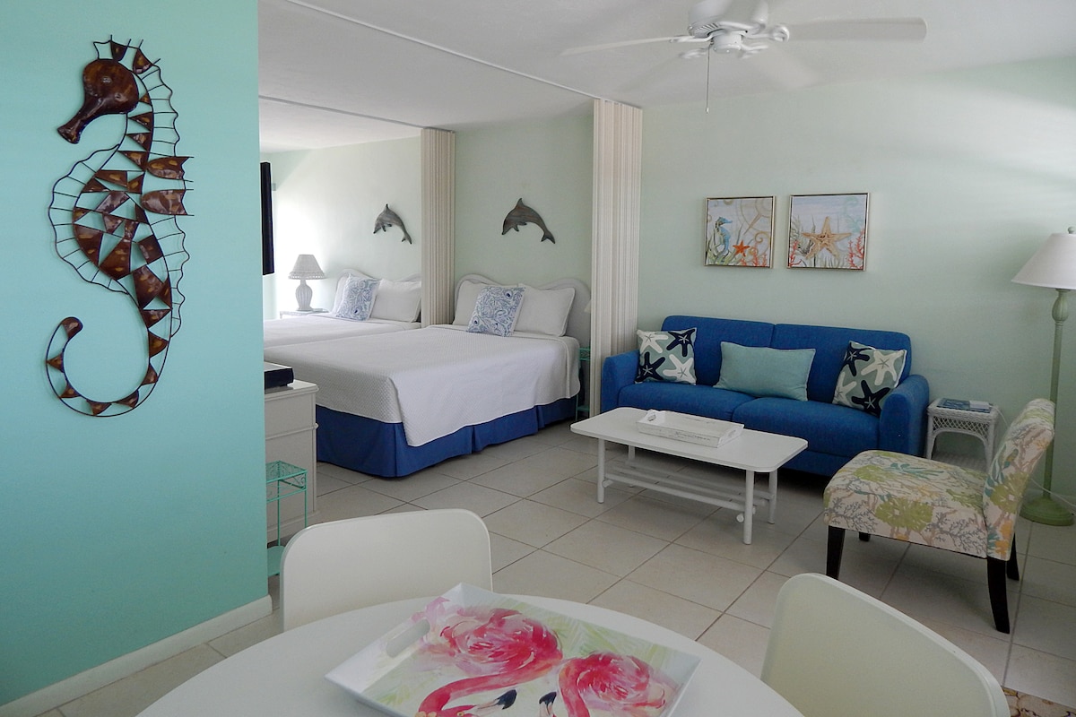 A bright studio condo is shown with two queen beds, separated by privacy screens. Tropical decor includes a seahorse sculpture and dolphin wall accents. A cozy blue couch is present, accompanied by a light-colored accent chair. A dining table with floral artwork completes the inviting space.
