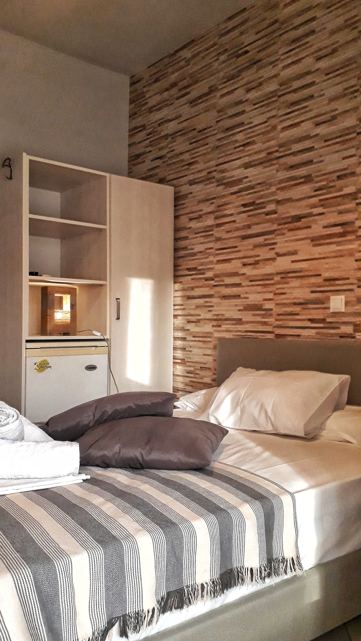 A cozy room is presented with a spacious bed dressed in soft linens and decorative pillows. A wardrobe and mini refrigerator are visible nearby. Textured stone-like wallpaper adds warmth, while natural light filters in, highlighting the inviting atmosphere of the space.