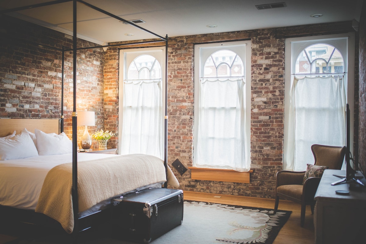 A spacious bedroom features exposed brick walls and large arched windows with light curtains. A comfortable bed is complemented by a decorative quilt. A vintage suitcase sits at the foot of the bed, and a cozy armchair is situated nearby, providing a relaxing seating area.