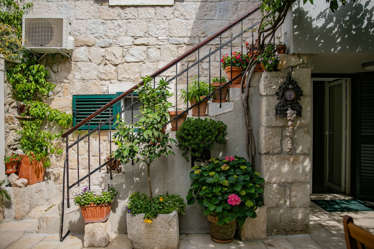 A charming stone staircase leads to the upper level of the building, adorned with pots of vibrant flowers and greenery. The natural elements complement the textured stone walls, creating a serene and inviting entryway. Sunlight illuminates the scene, enhancing the appeal of the outdoor area.