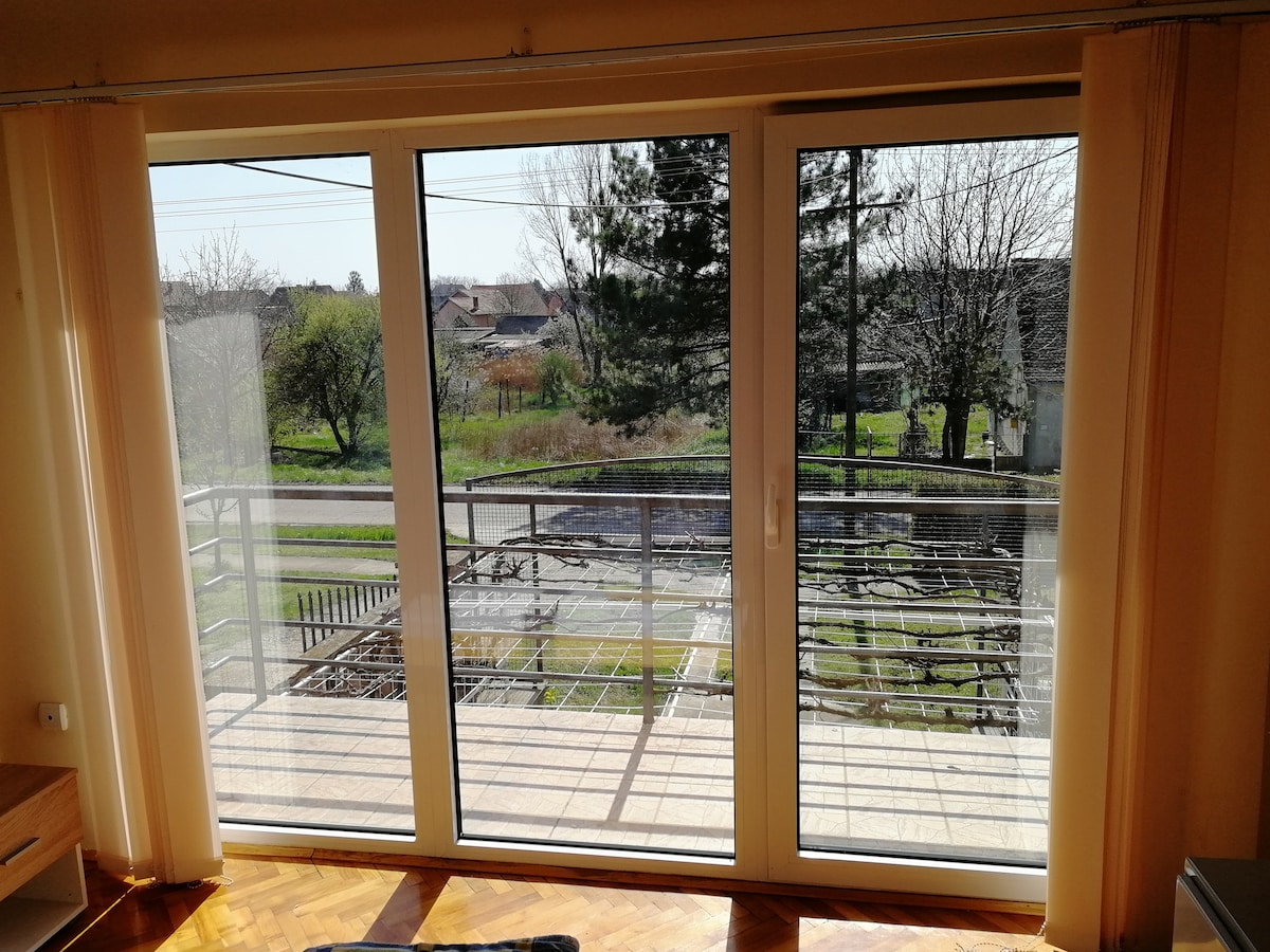 Large glass sliding doors provide a view of a green outdoor area with trees and a garden below. Natural light brightens the space, while a balcony railing defines the outdoor boundary. The flooring is composed of warm-toned wood, enhancing the serene ambiance.