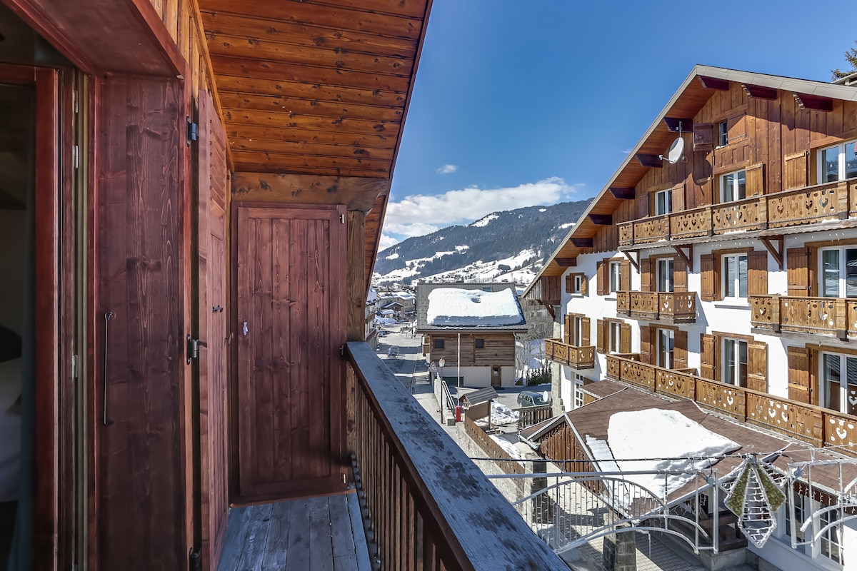 A wooden balcony provides a view of the surrounding chalet-style buildings and snowy mountains. The natural wood paneling complements the rustic aesthetic. Clear skies and distant slopes enhance the tranquil atmosphere, while walkways are visible below, indicating access to the nearby village.