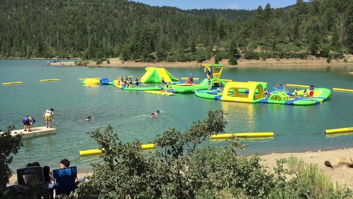 A vibrant floating water park is visible in the lake, featuring various inflatable slides and obstacles. Children can be seen swimming nearby, while a sandy area with greenery borders the water. The backdrop includes lush trees and mountains, creating a natural setting.