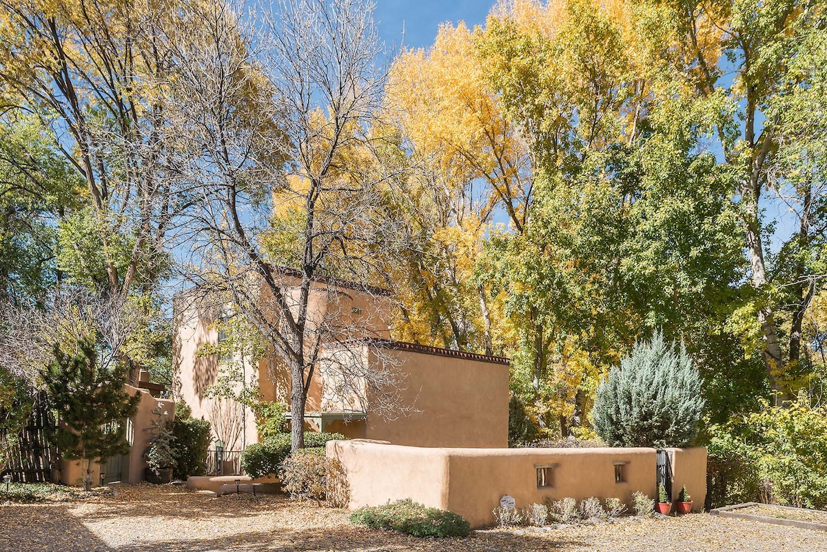 A secluded adobe building is surrounded by vibrant trees displaying autumn foliage. The warm earth-toned exterior complements the natural environment, while a patio area features colorful potted plants. A serene atmosphere is created by the interplay of light and shadow among the landscape.