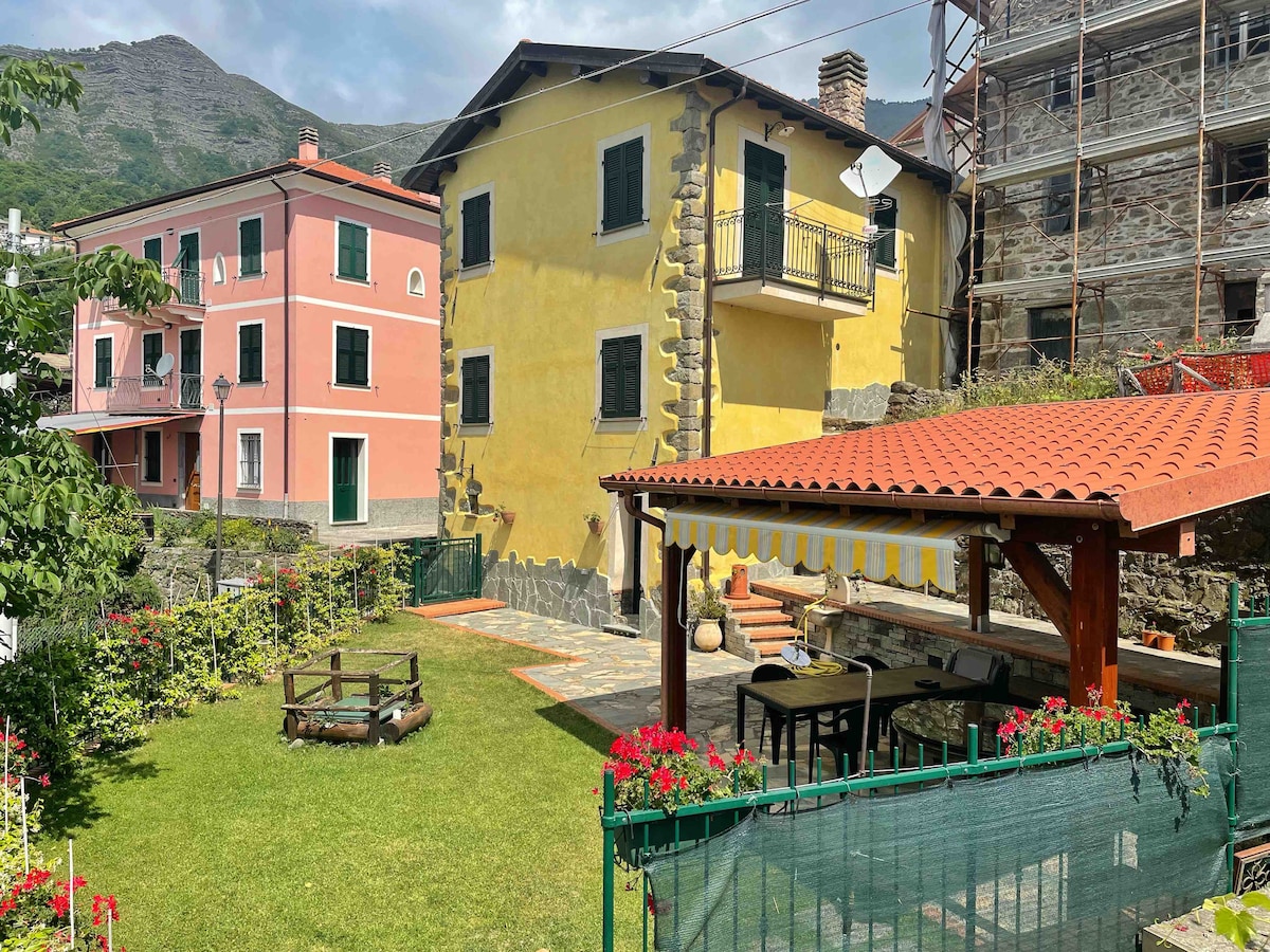 A charming exterior view of the house is presented, featuring a yellow façade and dark green shutters. A well-maintained garden is visible, complete with a patio area under a wooden pergola. Surrounding mountains and neighboring buildings are seen in the background.