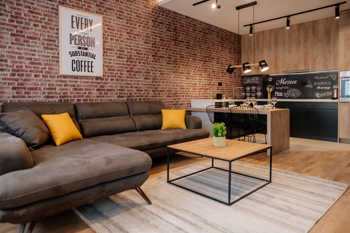A spacious living area features a modern brown sectional sofa adorned with bright yellow cushions. A wooden coffee table is positioned on a rug, enhancing the cozy ambiance. Open shelving and a menu board are visible behind a contemporary kitchen area with dark cabinetry.