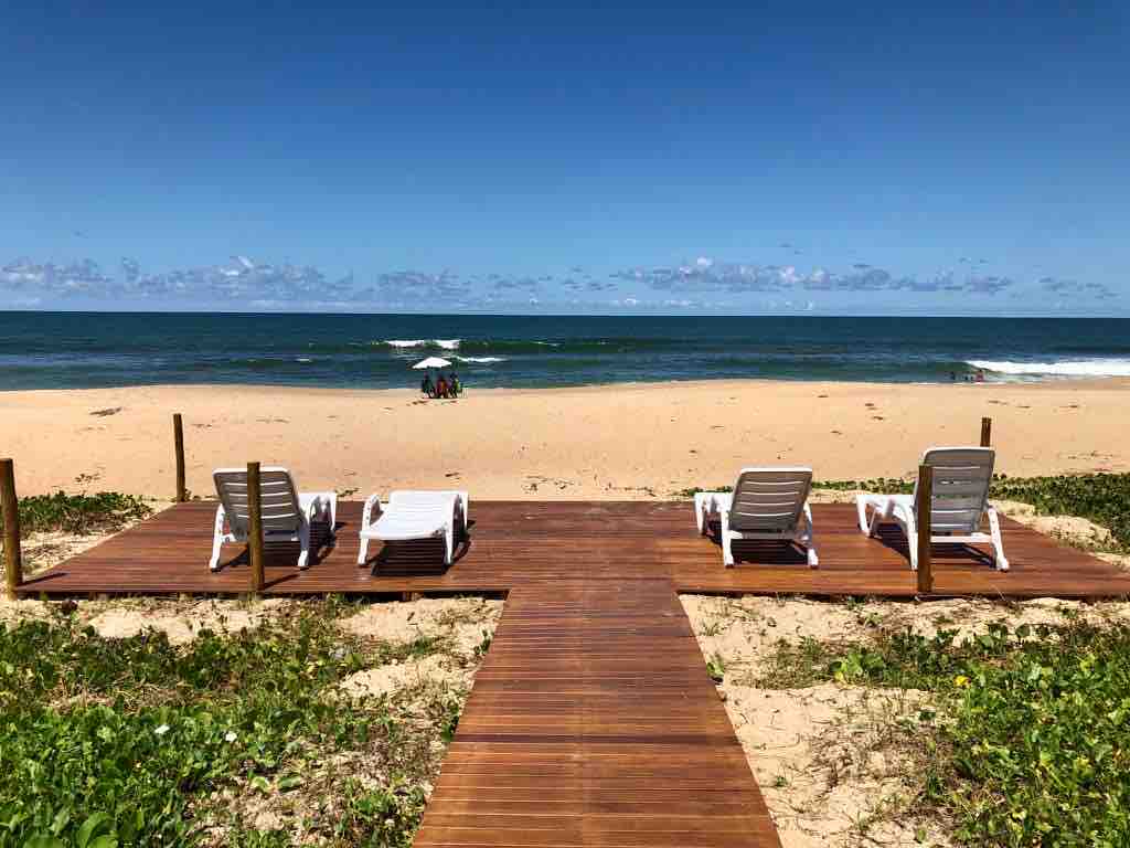 A wooden deck extends toward the sandy beach, providing a view of the ocean. Two white lounge chairs are positioned on the deck, inviting relaxation. The beach stretches into the distance, with waves gently lapping at the shore under a clear blue sky.