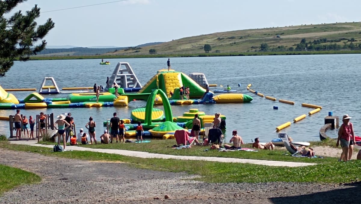 A waterpark is visible, featuring large inflatable structures in bright colors set against a lake backdrop. Individuals of various ages can be seen enjoying the attractions and relaxing on the sandy shore. Sun loungers are positioned along the beach, with grassy areas nearby.
