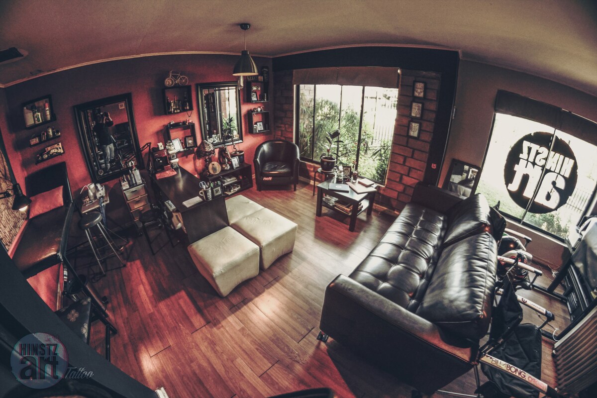 A stylish living room features a combination of dark wood and brick accents. Comfortable seating includes a large black sofa and two armchairs. Natural light filters through a large window, illuminating a small coffee table and decorative shelves filled with various items. A vibrant graphic is displayed on the window.