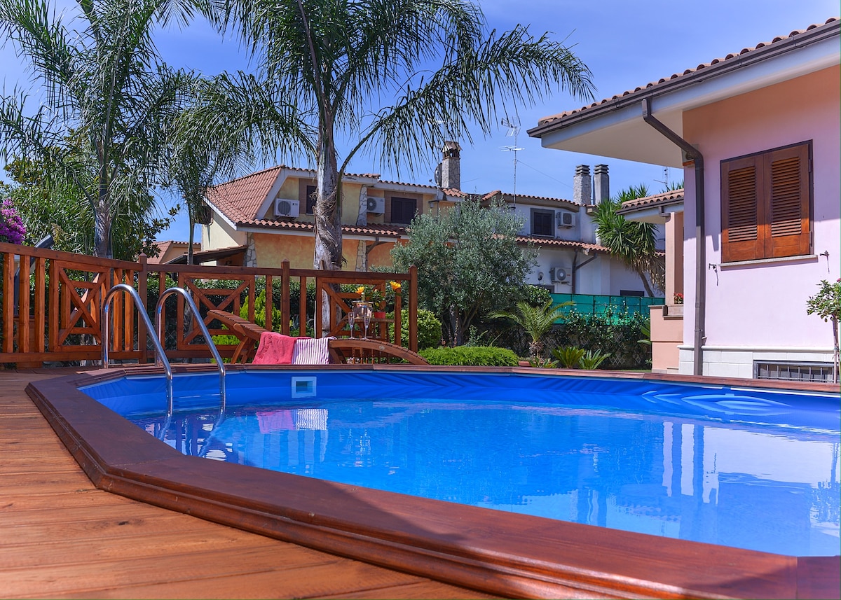 A private pool is surrounded by a wooden deck, featuring steps leading into the clear blue water. Lush palm trees provide shade, with a vibrant pink towel laid across the railing nearby. The villa's exterior is visible in the background, showcasing a harmonious residential setting.