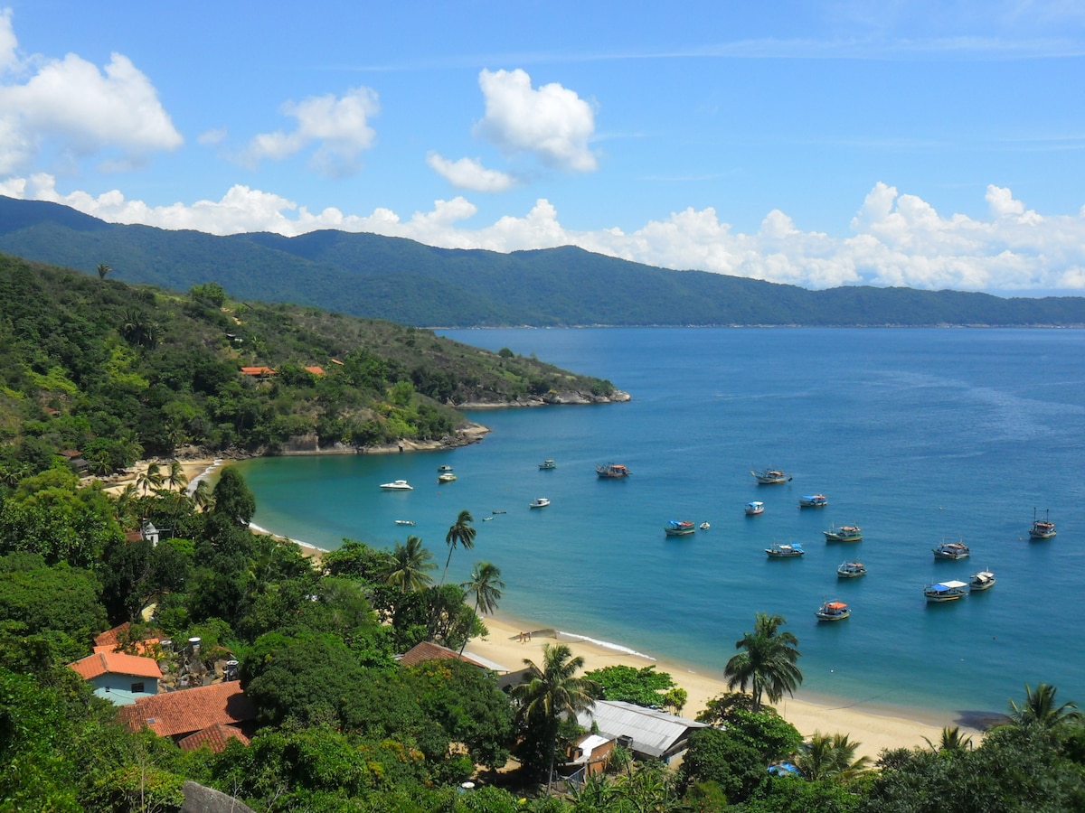 A serene beach is depicted, featuring calm waters and a sandy shoreline lined with lush greenery. Small boats are anchored in the clear blue water, with distant hills providing a tranquil backdrop under a bright sky with scattered clouds.
