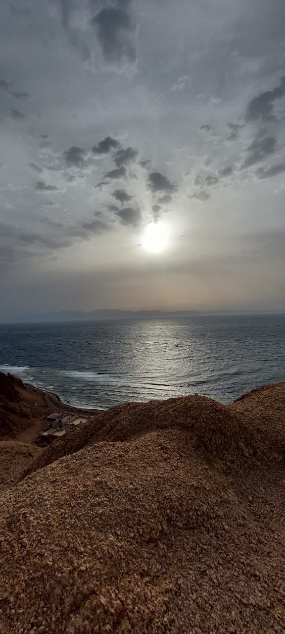 A coastal view captured at sunset, showing the sun partially obscured by clouds over the calm sea. Gentle waves reflect various shades of grey and soft silver on the water's surface, while the rugged coastline is visible in the foreground.