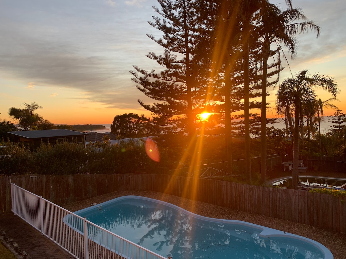 A serene sunset is captured over a tranquil pool, surrounded by lush trees. The warm glow of the sun filters through the branches, casting a gentle light on the landscape. A white fence encloses the pool area, enhancing the inviting atmosphere.