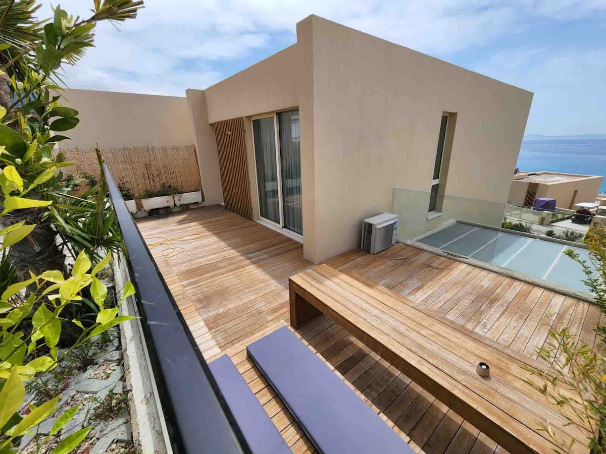 An outdoor terrace is shown with a wooden deck, featuring a sleek low table and a built-in bench. Large glass doors lead to the interior, while a mountain and sea view is visible in the distance, framed by potted greenery and natural elements.