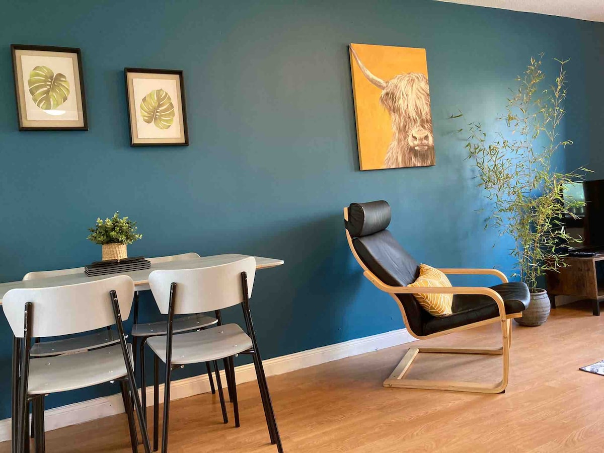 A dining area features a light wooden table accompanied by three chairs, set against a deep blue wall adorned with framed botanical prints. A decorative plant and a stylish black chair enhance the space, while a vibrant artwork of a highland cow adds a playful touch.