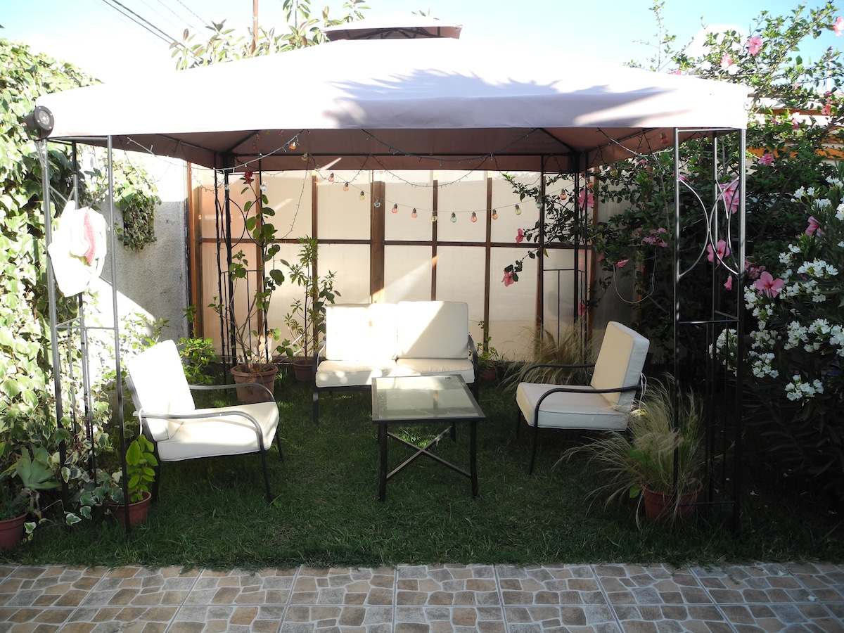 A shaded outdoor seating area is framed by greenery, featuring four white chairs surrounding a glass table. Colorful plants and flowers enhance the tranquil atmosphere, while soft lighting adds a welcoming touch. The grass-covered ground invites relaxation and leisure.