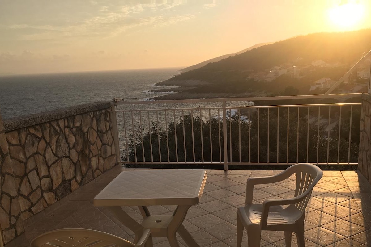 A balcony is displayed, featuring a white table and two plastic chairs. The expansive view overlooks the sea and coastline, with a sunset casting warm light over the landscape. The balcony is framed by a stone wall and has a railing for safety.