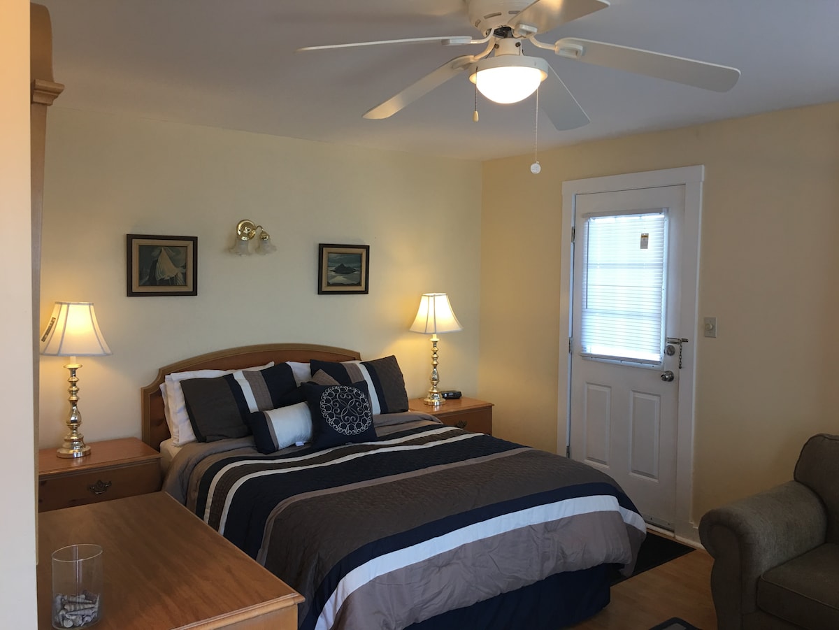 A cozy bedroom is furnished with a queen bed, complemented by bedside tables and lamps. A light ceiling fan circulates air above. The room includes a door leading to the exterior and features wall art that adds subtle color to the neutral walls.